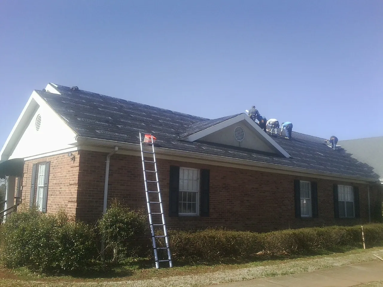 Crew tearing shingles off a brick ranch roof with a ladder set — Metal Roof Repair work by our crew in Erskine