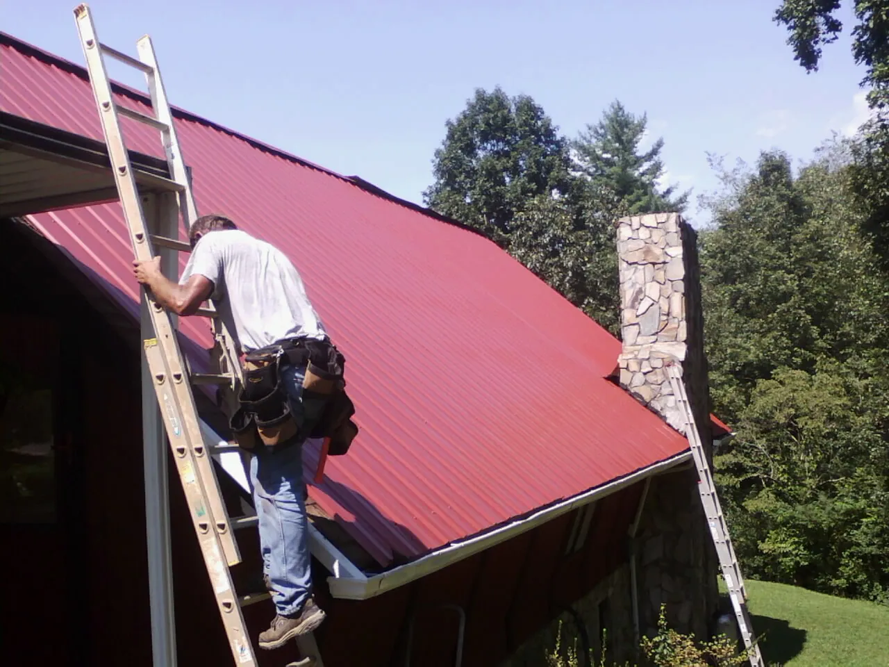Roofer on a ladder beside a red metal roof and stone chimney — Soffit & Fascia Repair by Roofing Brothers in Erskine